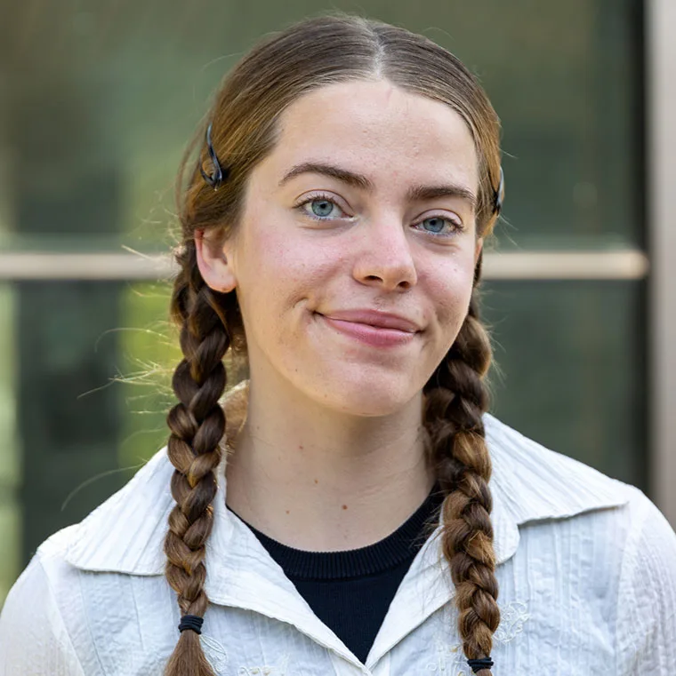 Person with long hair in braids, wearing a white shirt and a black top, smiles slightly in front of a blurred background.