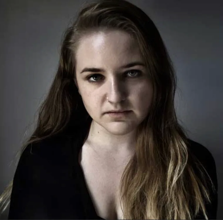Person with long hair looking directly at the camera against a dark background, wearing a black top.