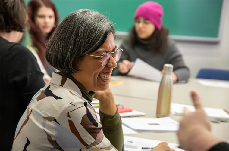 Person with glasses smiling at a table with others talking and taking notes. One person wears a pink hat. Background features a chalkboard.