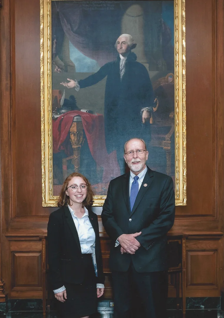 Two people stand in front of a large portrait in an ornate frame, set within a wood-paneled room.