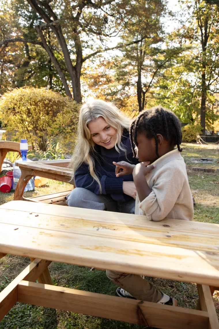 Person smiling and interacting with a child outdoors at a picnic table with trees and autumn leaves in the background.