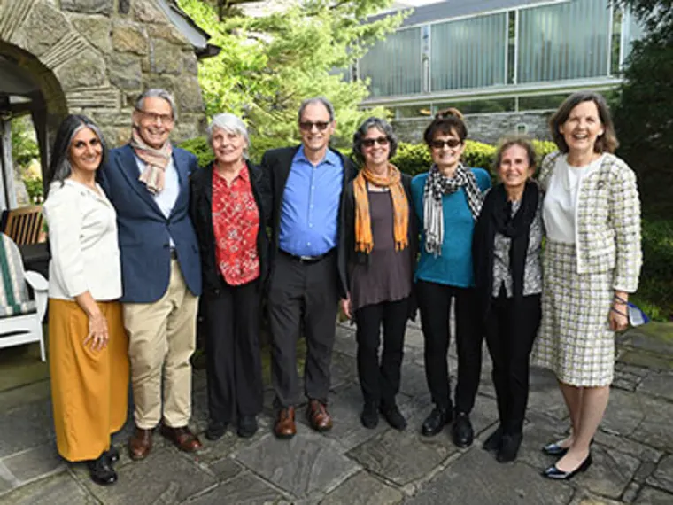 Eight people standing together outside, smiling, in front of a stone building with greenery around.