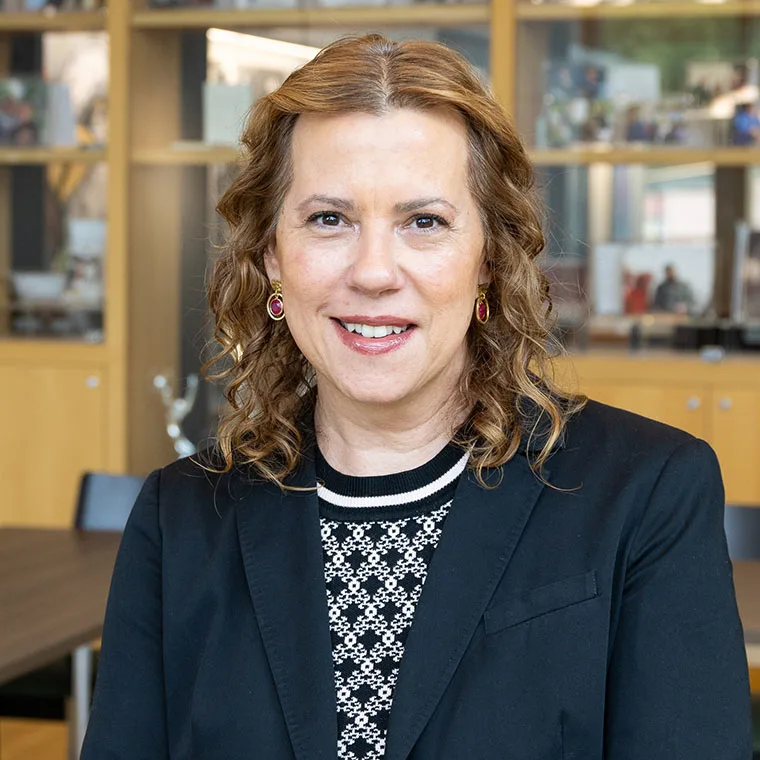 Person with curly hair wearing a black blazer and patterned shirt in an office setting, smiling at the camera. Shelves in the background.