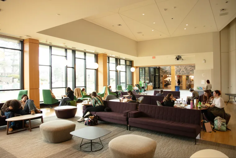 People sitting on couches and chairs in a spacious, sunlit lounge area with large windows and round lights.