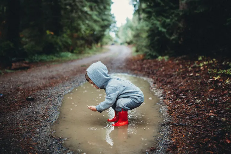 Person in a gray hoodie and red boots crouches by a puddle on a forest path, surrounded by trees.
