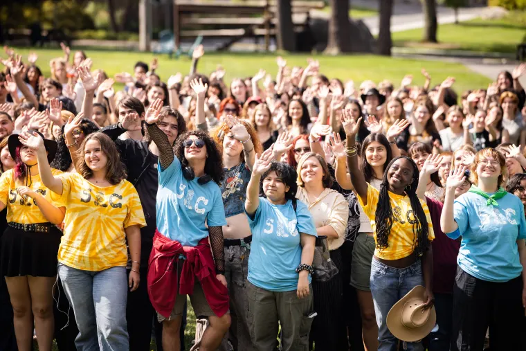 A large group of people outdoors in colorful shirts smiling and waving at the camera, with trees and grass in the background.