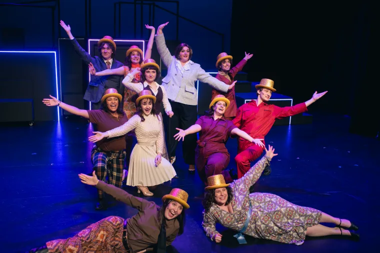 A group of performers on stage wearing gold hats and colorful outfits, striking cheerful poses with arms extended.