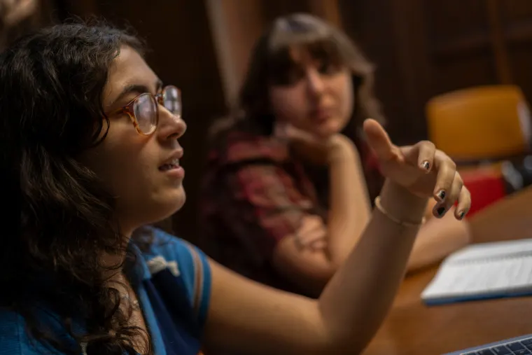 Person in glasses gestures while speaking at a table. Another person listens in the background with a notebook nearby.