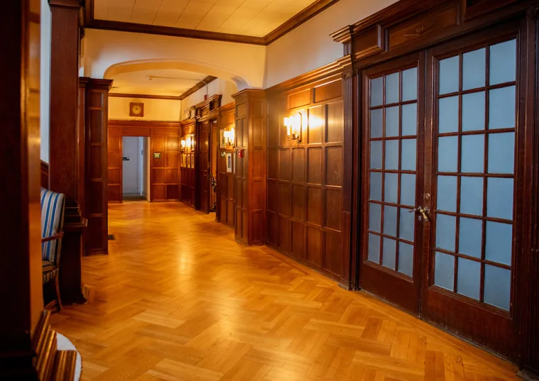 Wood-paneled hallway with parquet flooring, well-lit by wall sconces. Glass-paned doors on the right and a corridor leading further inside.