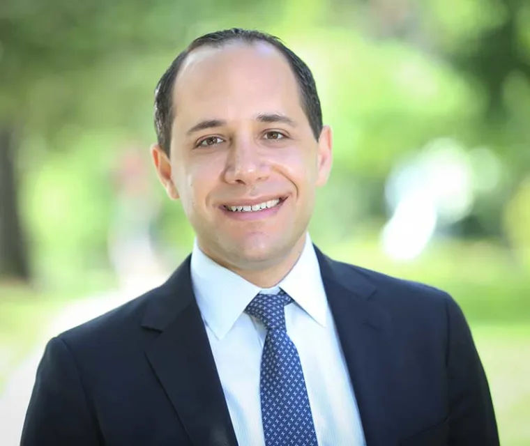 Person in a suit with a tie smiling outdoors, with a blurred green background.