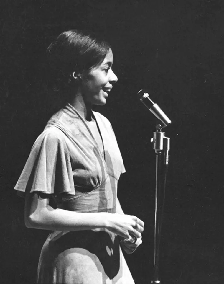 Person standing at a microphone, smiling, with a dark background, wearing a dress with short sleeves.