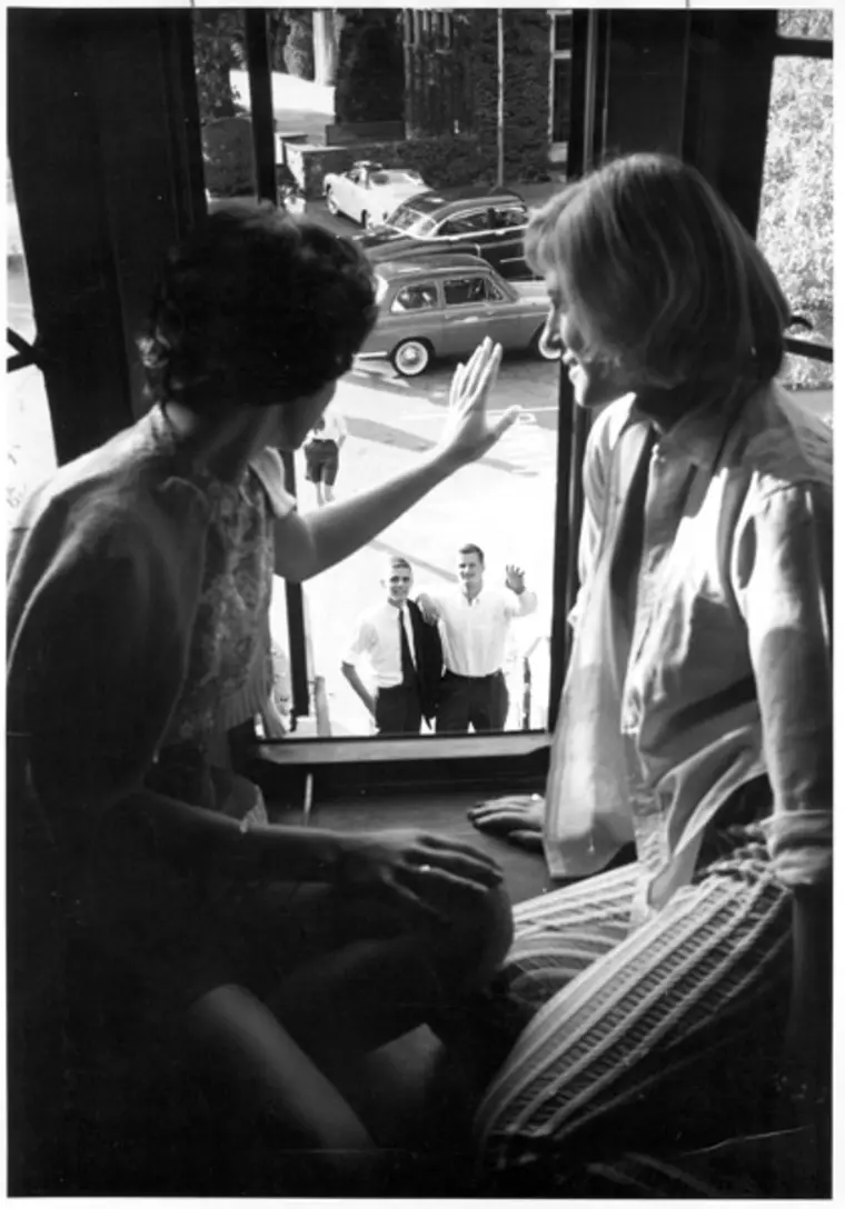 Two people sit by a window, one waving to two individuals outside. Several vintage cars are parked on the street below.