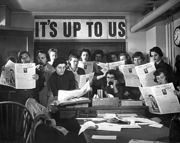 Group of people reading newspapers in an office under a sign that says "It's Up to Us." One person is talking on the phone.