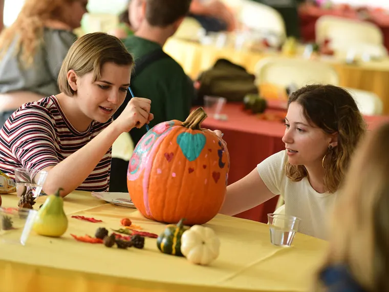 Two people decorate an orange pumpkin with colorful designs at a table with small gourds and art supplies.