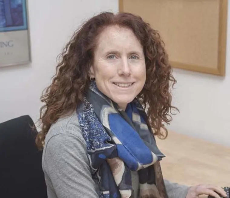 Person with curly hair sitting at a desk, wearing a patterned scarf, smiling at the camera. A corkboard is in the background.