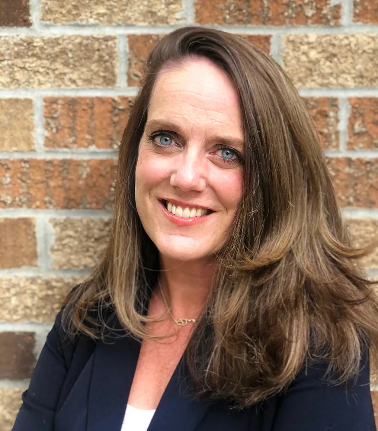 Smiling person with long brown hair stands in front of a brick wall, wearing a dark blazer and a necklace.