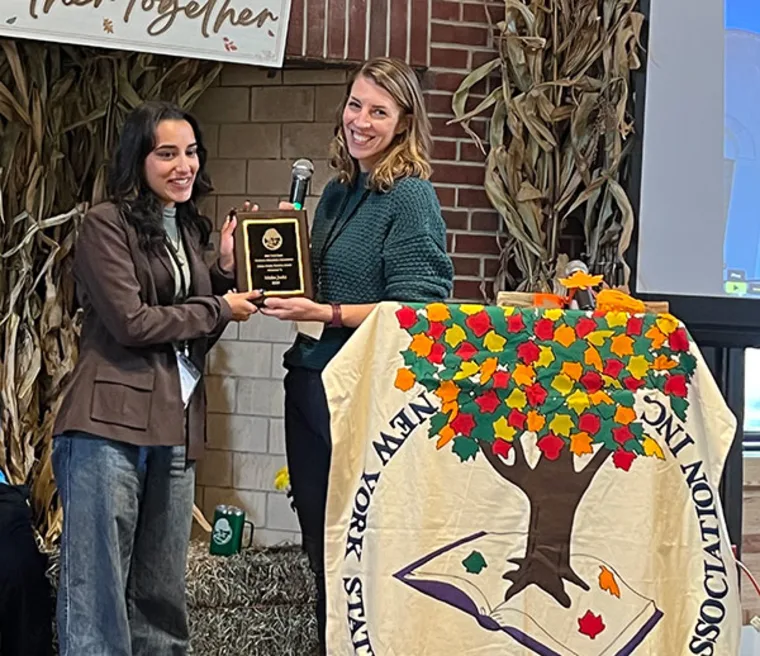 Two individuals are smiling and holding a plaque. They stand behind a podium covered with a cloth featuring a tree and colorful leaves.