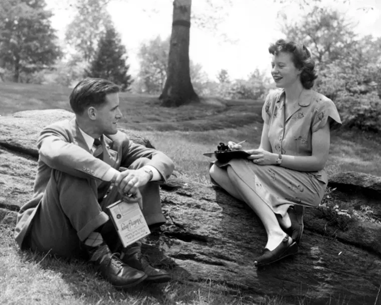 Two people sitting on a rock in a park, one holds a bag, the other writes on a notepad. Trees and grass are visible in the background.