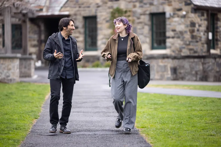 Two people walking on a path, engaged in conversation, with a stone building in the background.