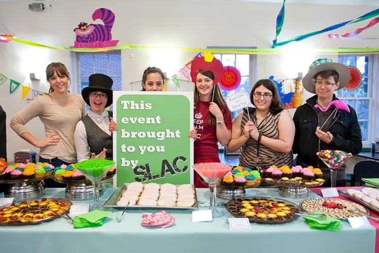Group of people behind a table of colorful desserts with a sign saying "This event brought to you by SLAC," decorated with streamers.