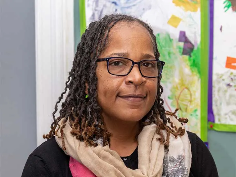 Person with glasses and locs, wearing a scarf, stands in front of colorful artwork on the wall.