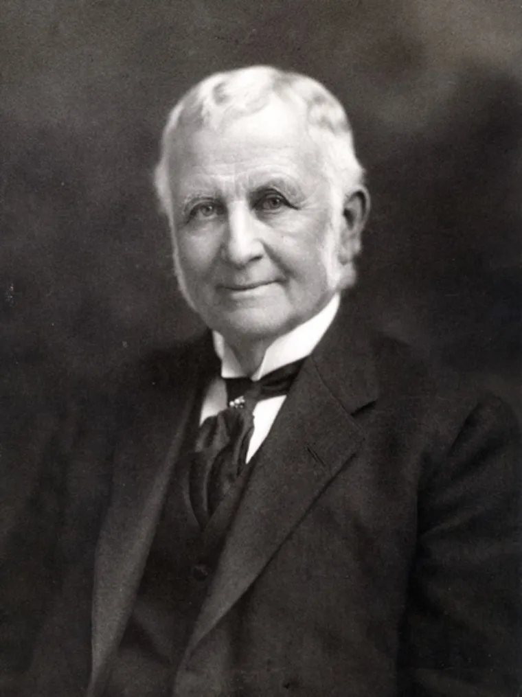 A person with short, white hair and sideburns, wearing a suit and tie, in a sepia-toned portrait with a neutral expression.