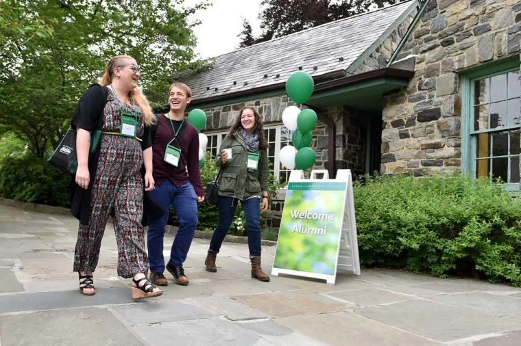 Three people with lanyards walk past a "Welcome Alumni" sign outside a stone building with green and white balloons.