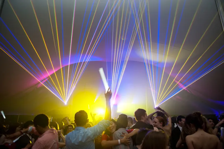 Crowded dance floor under colorful laser lights, with people holding glow sticks and enjoying the vibrant party atmosphere.