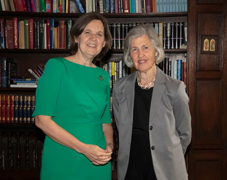 Two people smiling, standing side by side in front of a bookshelf filled with various books. One wears green, the other grey.