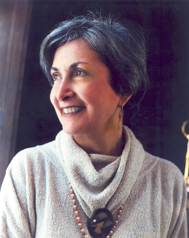 Person with short gray hair smiling and looking to the side, wearing a light sweater and a pendant necklace.