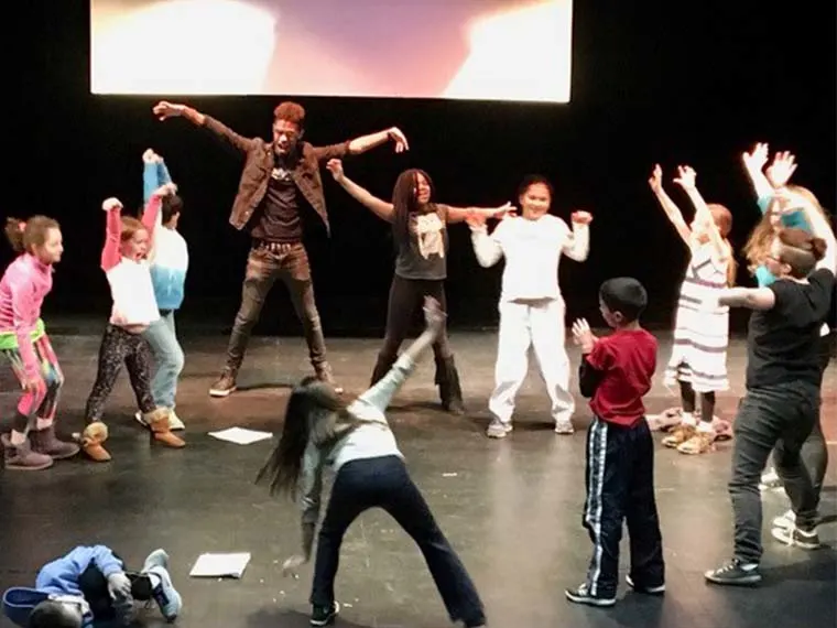 A group of children and an adult on a stage, energetically performing a dance routine with arms raised and wide stances.
