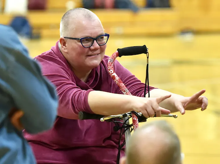 Person with short hair wearing glasses and a maroon shirt, sitting on a mobility scooter, gesturing with both hands in an indoor setting.