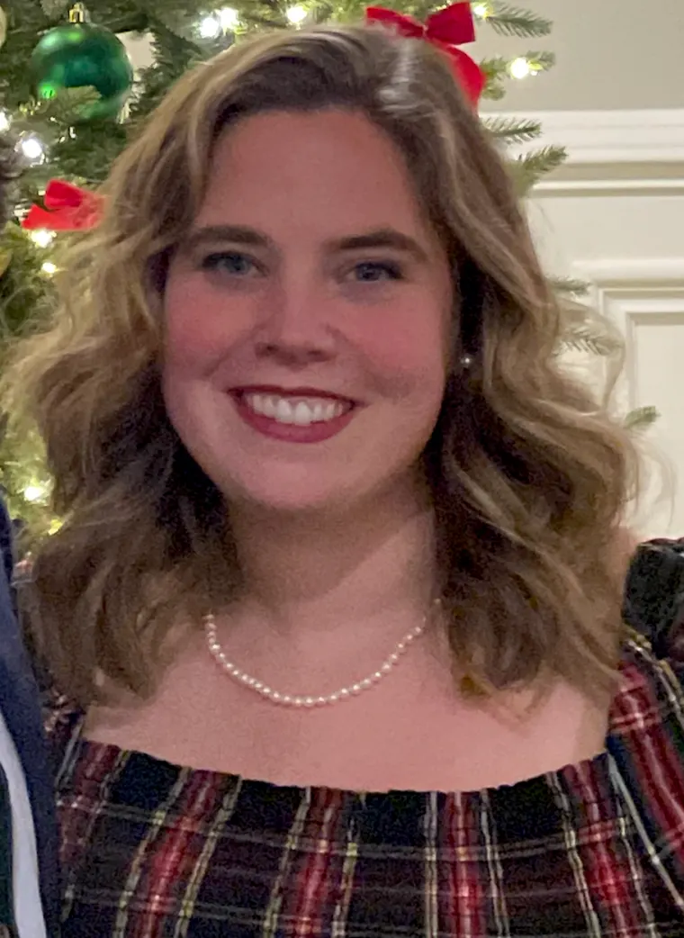 Person smiling in front of a Christmas tree, wearing a plaid outfit and a pearl necklace, with ornaments visible in the background.