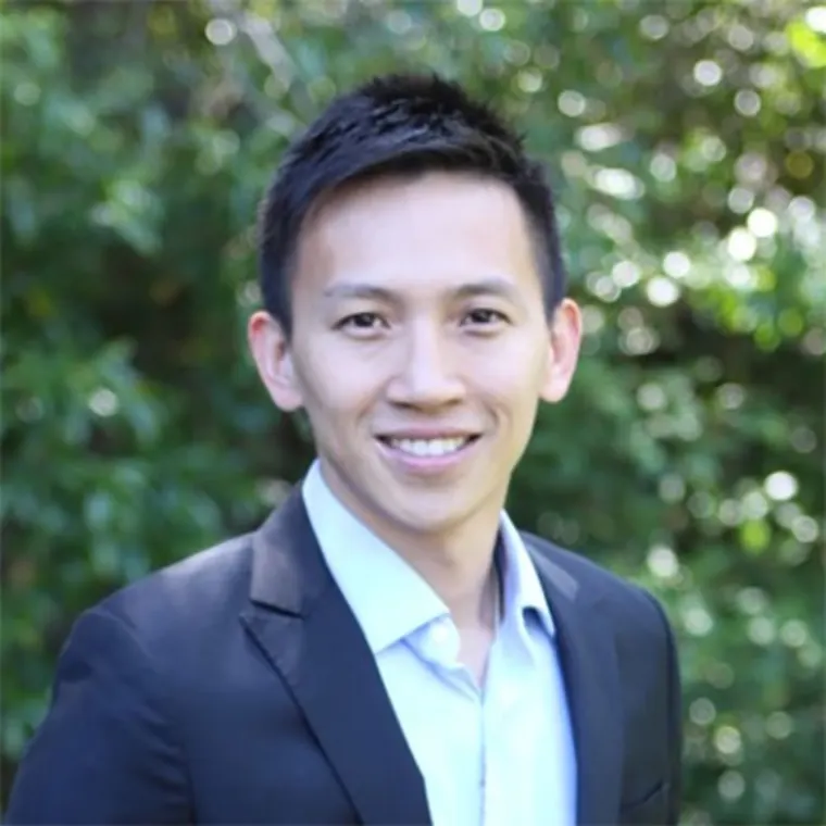 Person smiling, wearing a dark blazer and light shirt, standing outdoors with green foliage in the background.