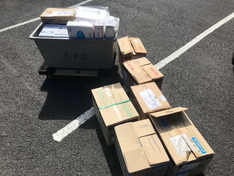 Multiple cardboard boxes and a grey bin filled with packages are stacked on a parking lot with white painted lines.