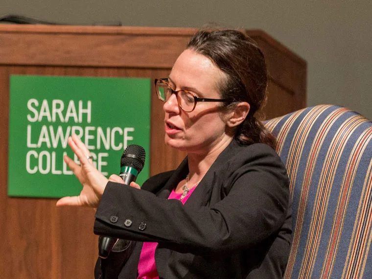 Person speaking into a microphone, seated in a striped chair, with a Sarah Lawrence College sign in the background.