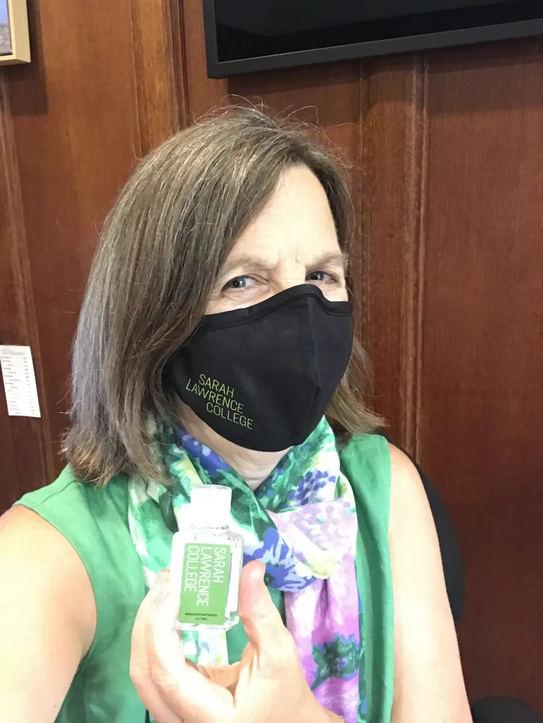 Person wearing a mask with "Sarah Lawrence College" text, holding a bottle with the same label, standing in front of a wooden wall.