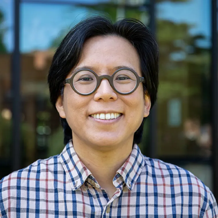 Person with short dark hair, wearing round glasses and a plaid shirt, smiling outdoors with blurred background.