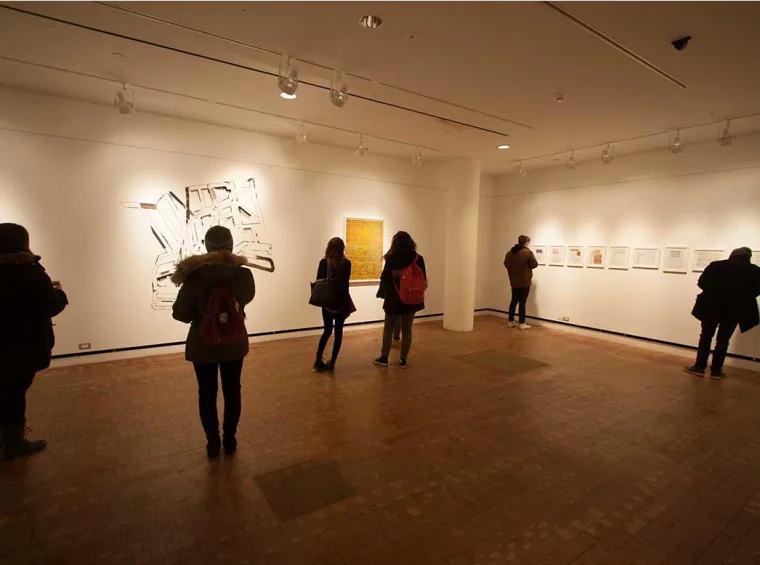 Visitors in a dimly lit gallery viewing abstract and framed art on white walls, some wearing backpacks and coats.