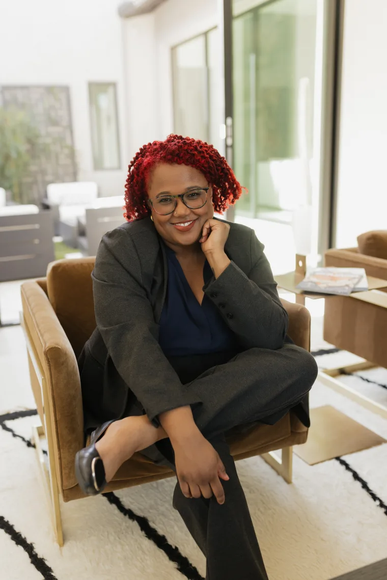 A person with red curly hair and glasses sits cross-legged on a chair, wearing a suit and smiling in a modern, well-lit room.
