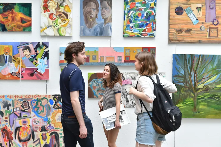 Three people stand talking and smiling in front of a wall filled with colorful paintings and artwork.