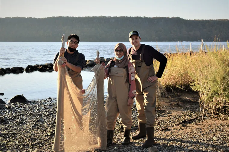 Three people in waders stand on a rocky shore holding nets beside a calm body of water, with trees in the background.