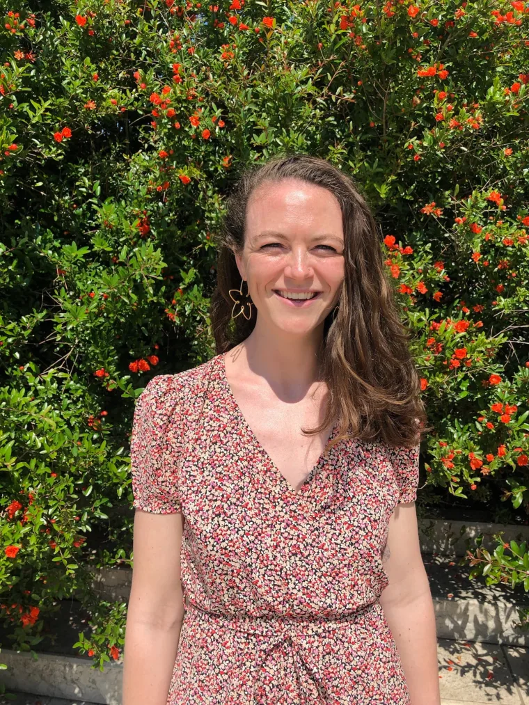 Person in a floral dress stands smiling in front of a bush with small red flowers on a sunny day.