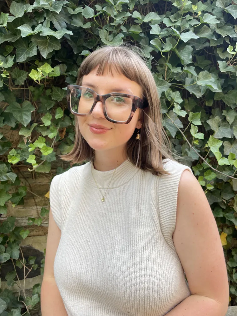 Person wearing large glasses and a sleeveless top, standing in front of a wall covered in green ivy leaves.