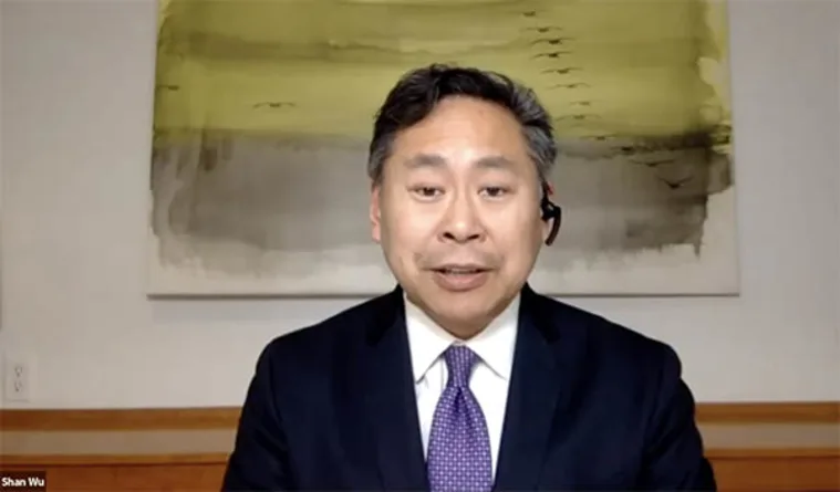 A person wearing a suit and tie with a headset speaks in front of an abstract painting.