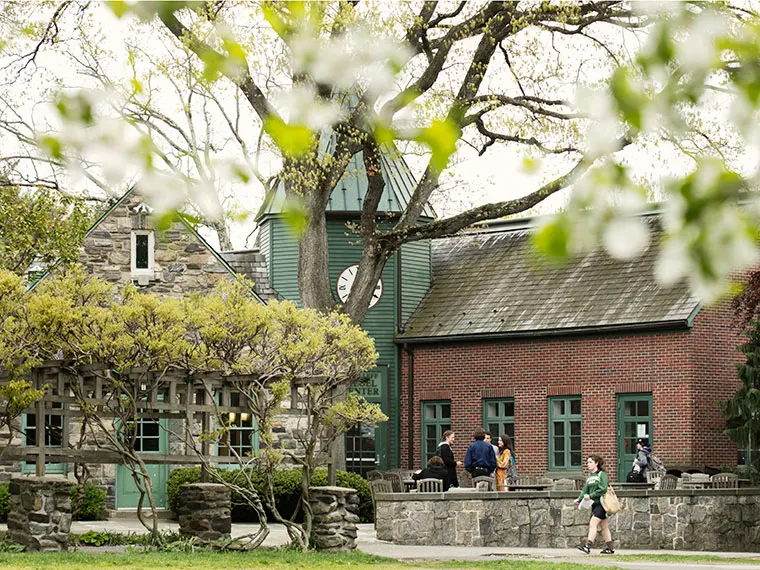 People walk near a brick building with a green clock tower, surrounded by trees and spring blossoms.