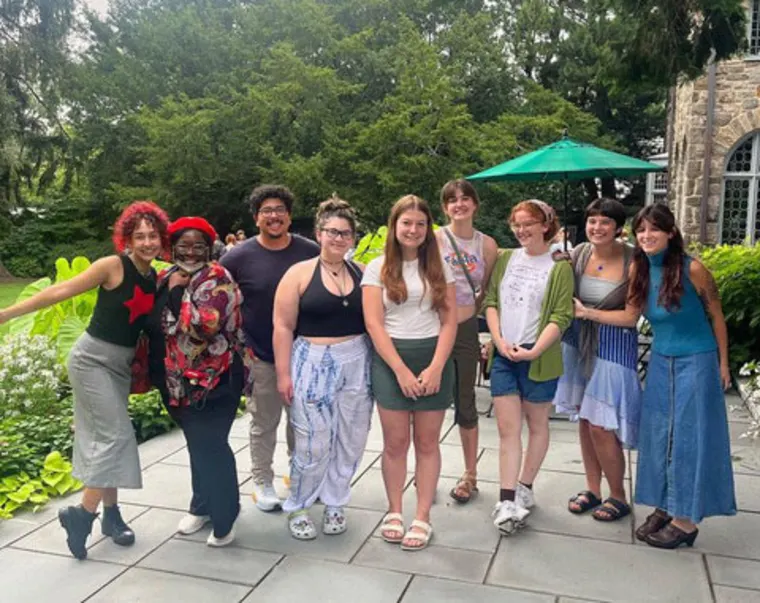 A group of nine people smiling outdoors on a patio with trees and a green umbrella in the background.