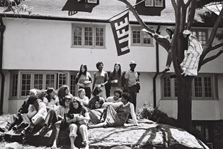 A group of people gather on and around a large rock in front of a building. A "LIFE" flag hangs nearby, and one person is perched on a tree branch.