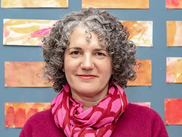 Person with curly gray hair wearing a pink scarf and sweater, standing against a colorful abstract background.