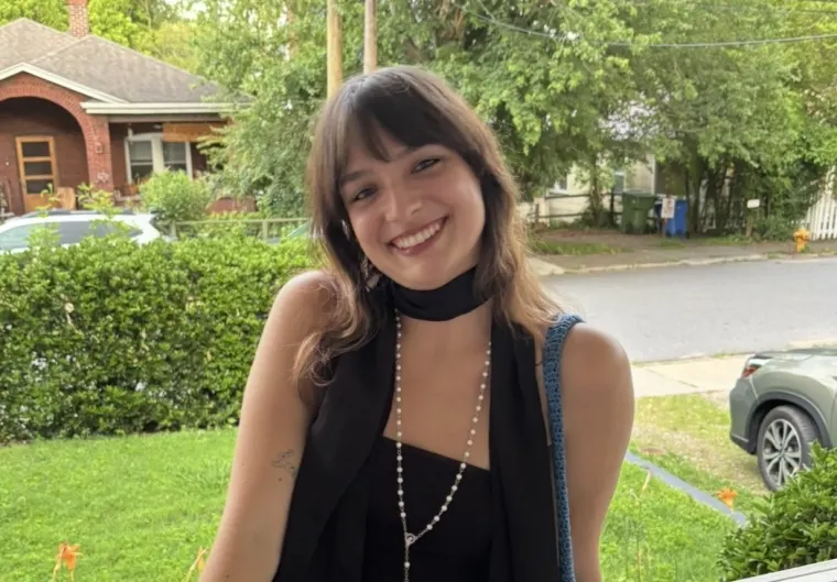 Person smiling outside with short hair and fringe, wearing a black top and scarf, beaded necklace, with a suburban house and trees in the background.